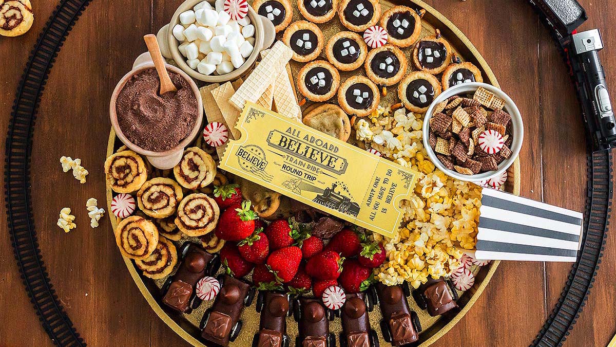 A round tray filled with various snacks including strawberries, popcorn, chocolate treats, cookies, marshmallows, and candies, with a yellow train ticket and striped popcorn bag in the center.