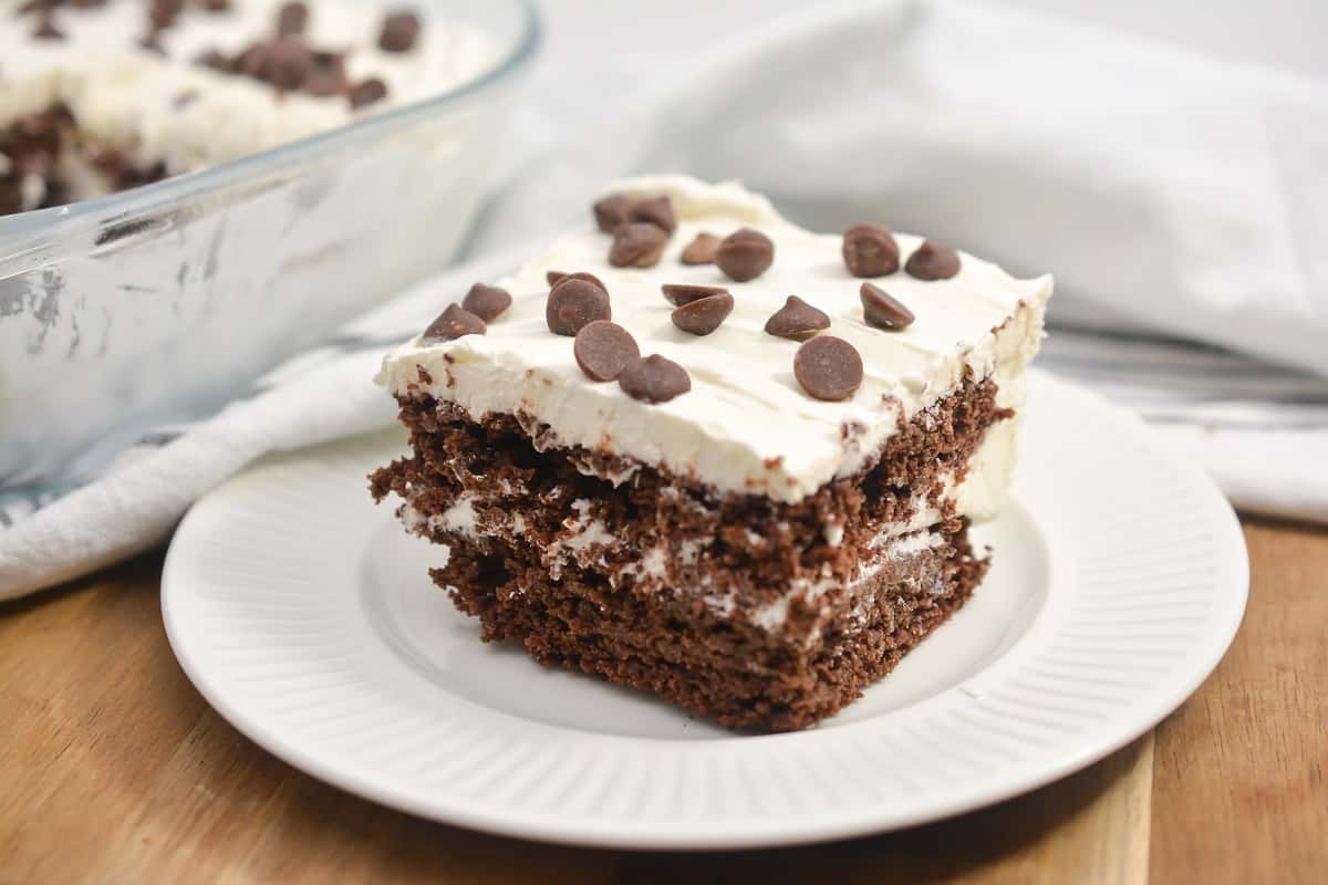 A slice of chocolate layer cake with white frosting and chocolate chips on top, served on a white plate with a glass baking dish in the background.