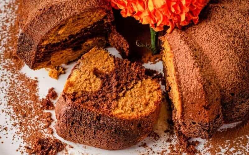 A sliced marble bundt cake with chocolate and vanilla swirls, dusted with cocoa powder and decorated with an orange flower.