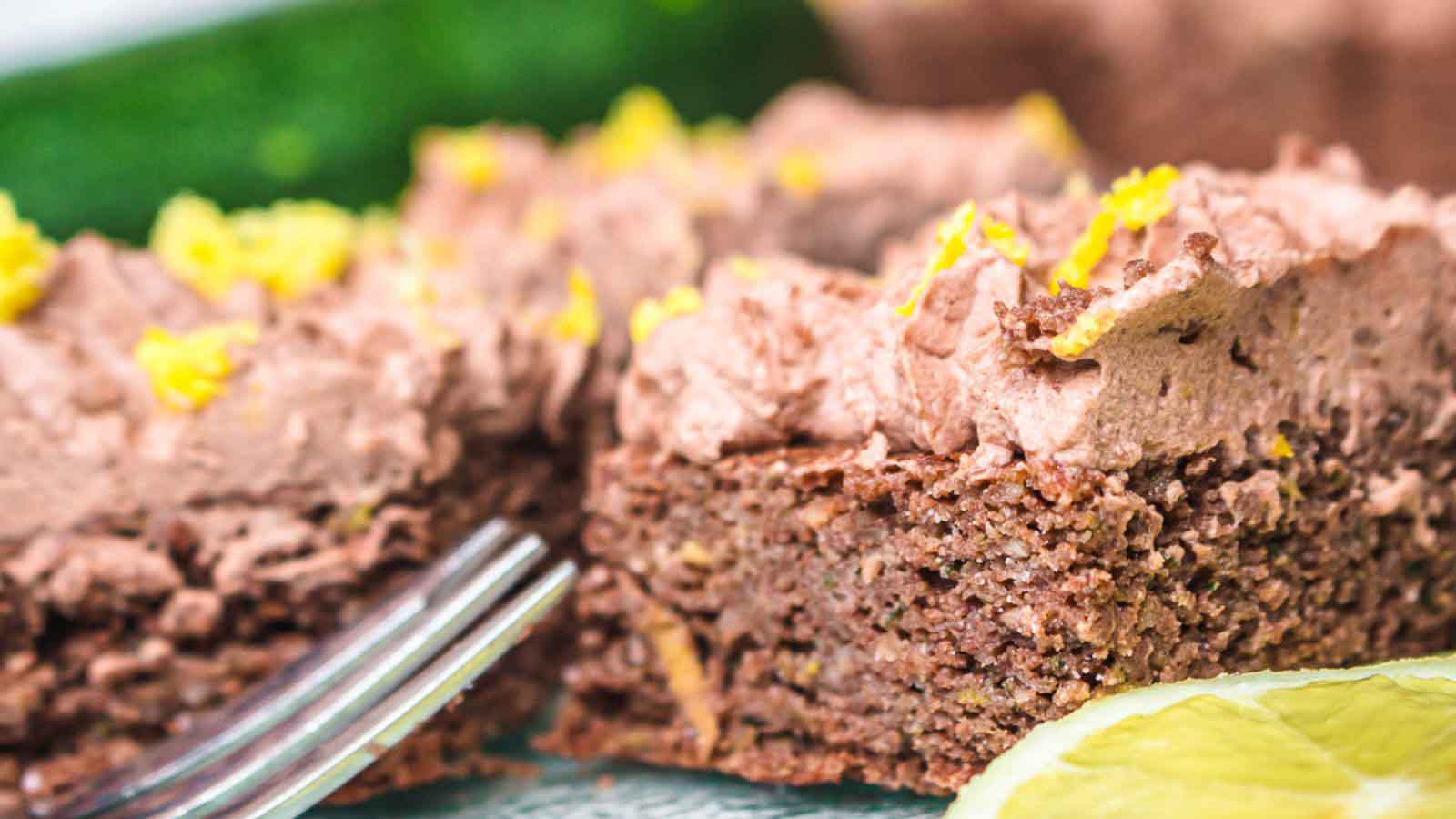 A close-up of chocolate frosted zucchini cake topped with lemon zest, with a fork and a slice of lemon in the foreground.