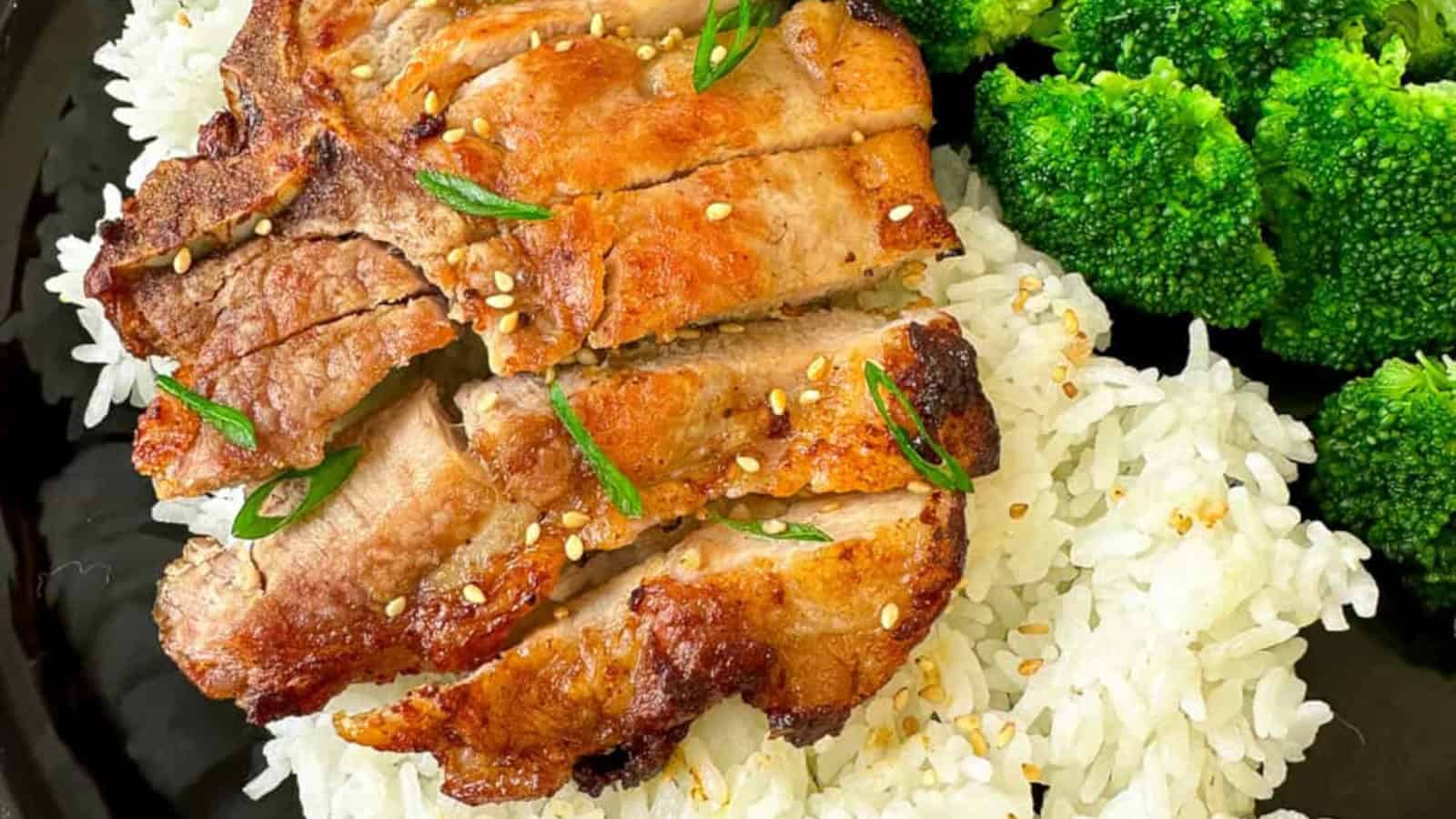 Grilled sliced pork with sesame seeds and green onions on white rice, served with steamed broccoli on a black plate.