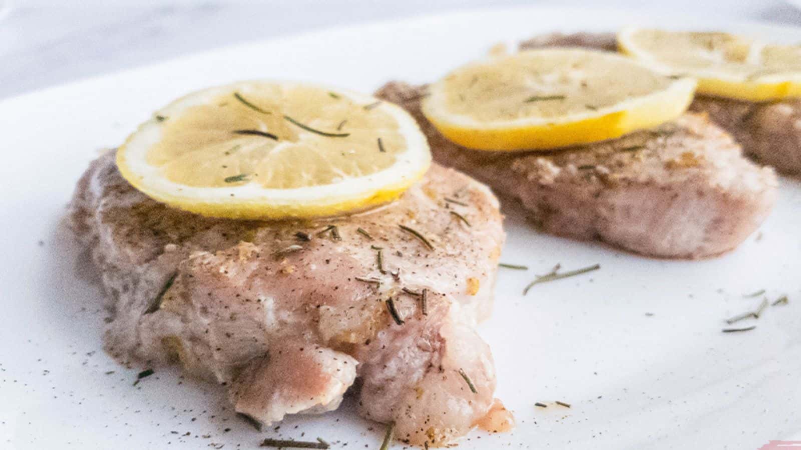 Three cooked pork chops garnished with lemon slices and sprinkled herbs, served on a white plate.