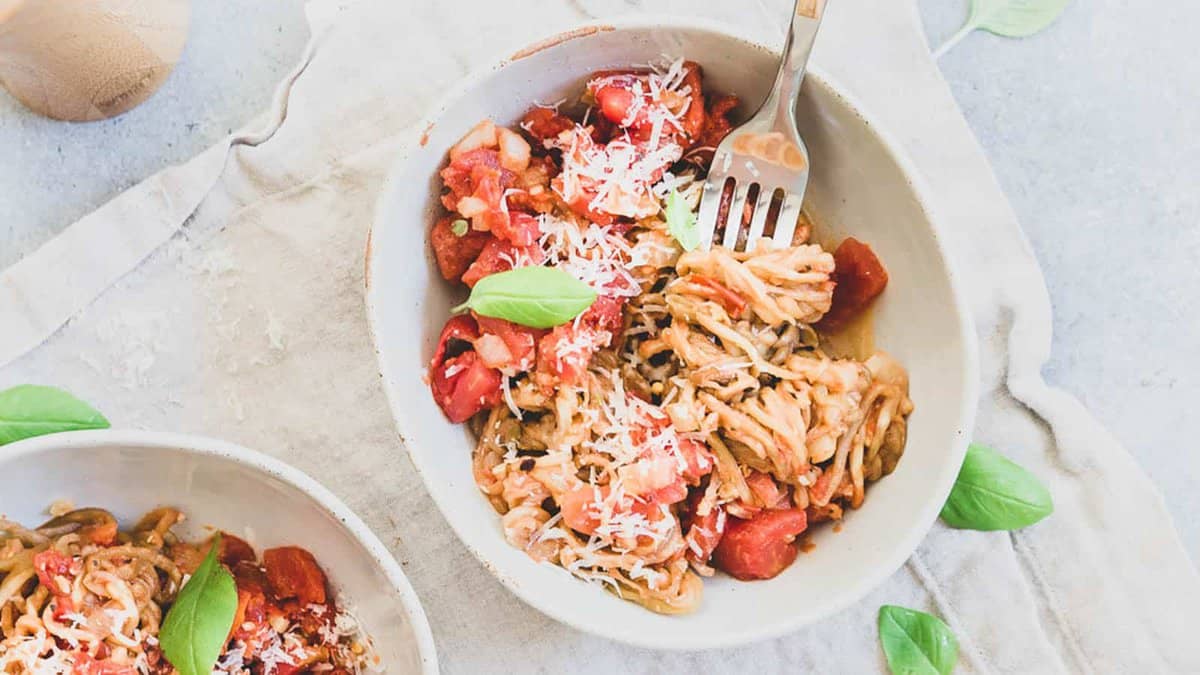 A bowl of spaghetti with tomato sauce, grated cheese, and fresh basil leaves on a light cloth napkin, with a fork resting in the bowl.