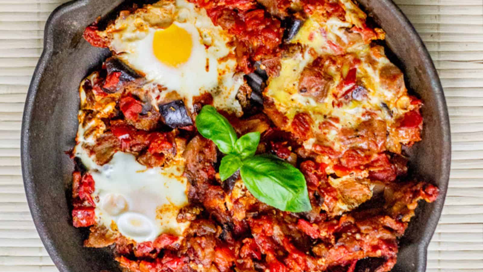 A skillet filled with shakshuka, containing eggs poached in a tomato and vegetable sauce, garnished with fresh basil leaves.