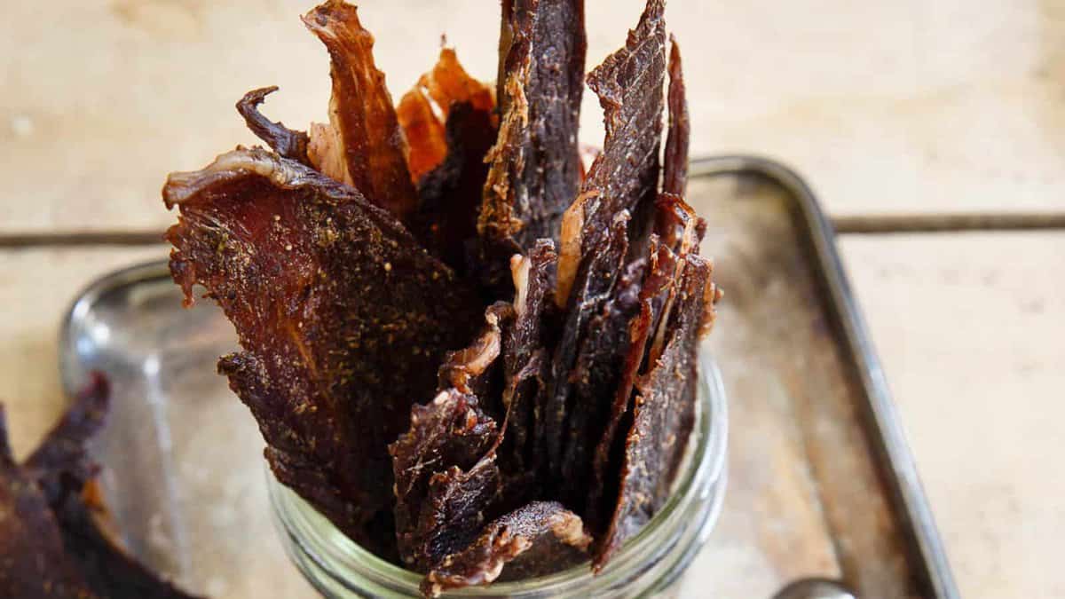 Several pieces of dark, dried beef jerky are arranged upright in a glass jar on a metal tray atop a wooden surface.