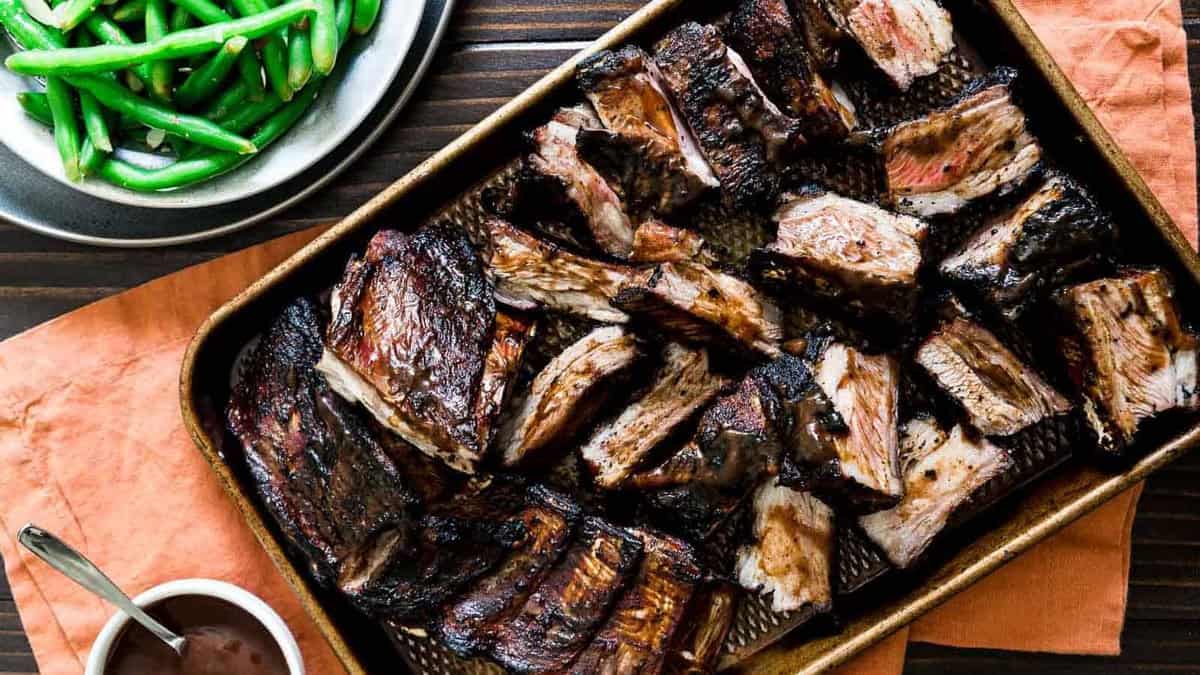 A tray of cooked, sliced beef short ribs next to a bowl of green beans and a small dish of sauce, set on a wooden table with an orange napkin.