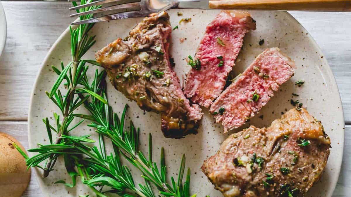 A plate with sliced, medium-rare steak garnished with fresh rosemary, next to a fork and knife.