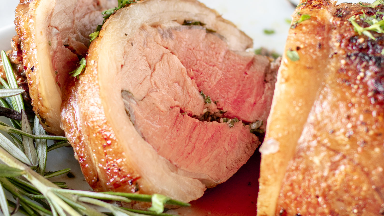 Close-up of sliced, cooked rolled meat, showing a pink center and seasoned with herbs, with sprigs of fresh rosemary on the side.