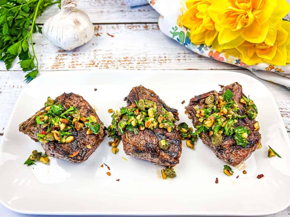 Three grilled lamb chops topped with a green herb and nut garnish, served on a white rectangular plate, with garlic, parsley, and yellow flowers in the background.