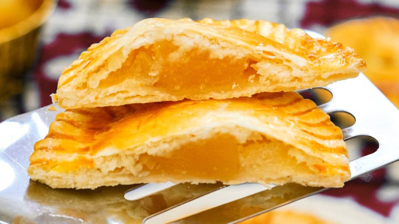 A close-up of a metal spatula holding two halves of a golden-brown pastry filled with yellow fruit filling, possibly pineapple jam.