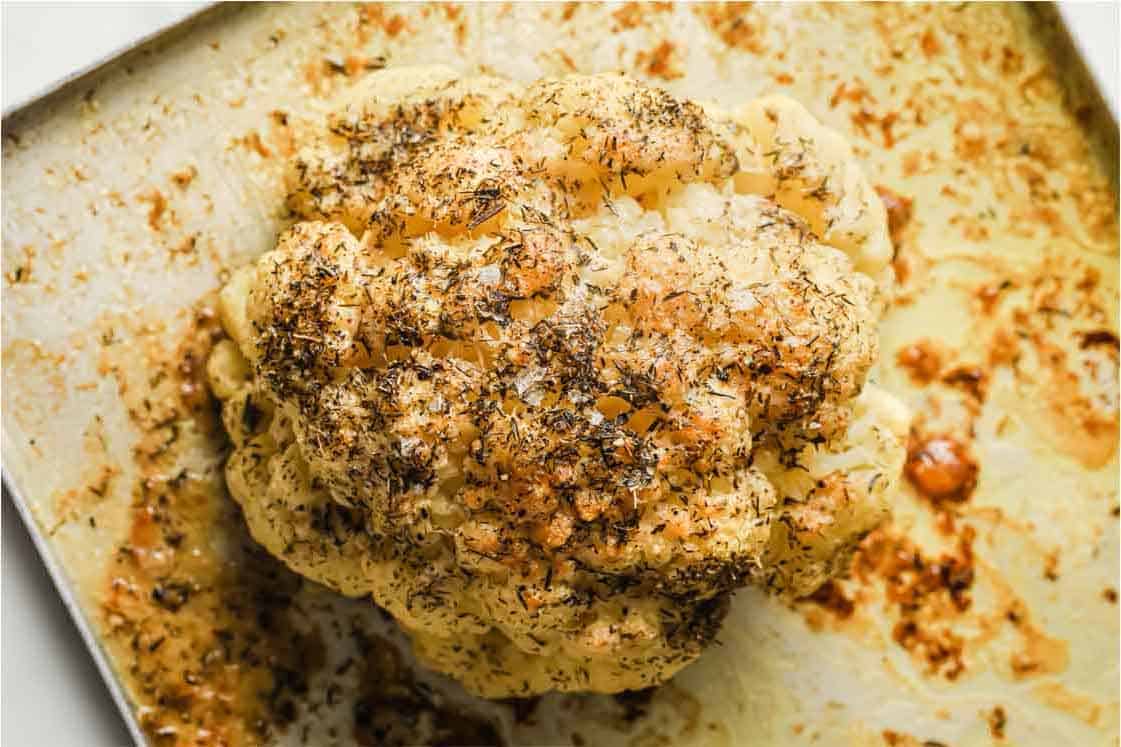 A whole roasted cauliflower head with spices and herbs sits on a baking sheet.
