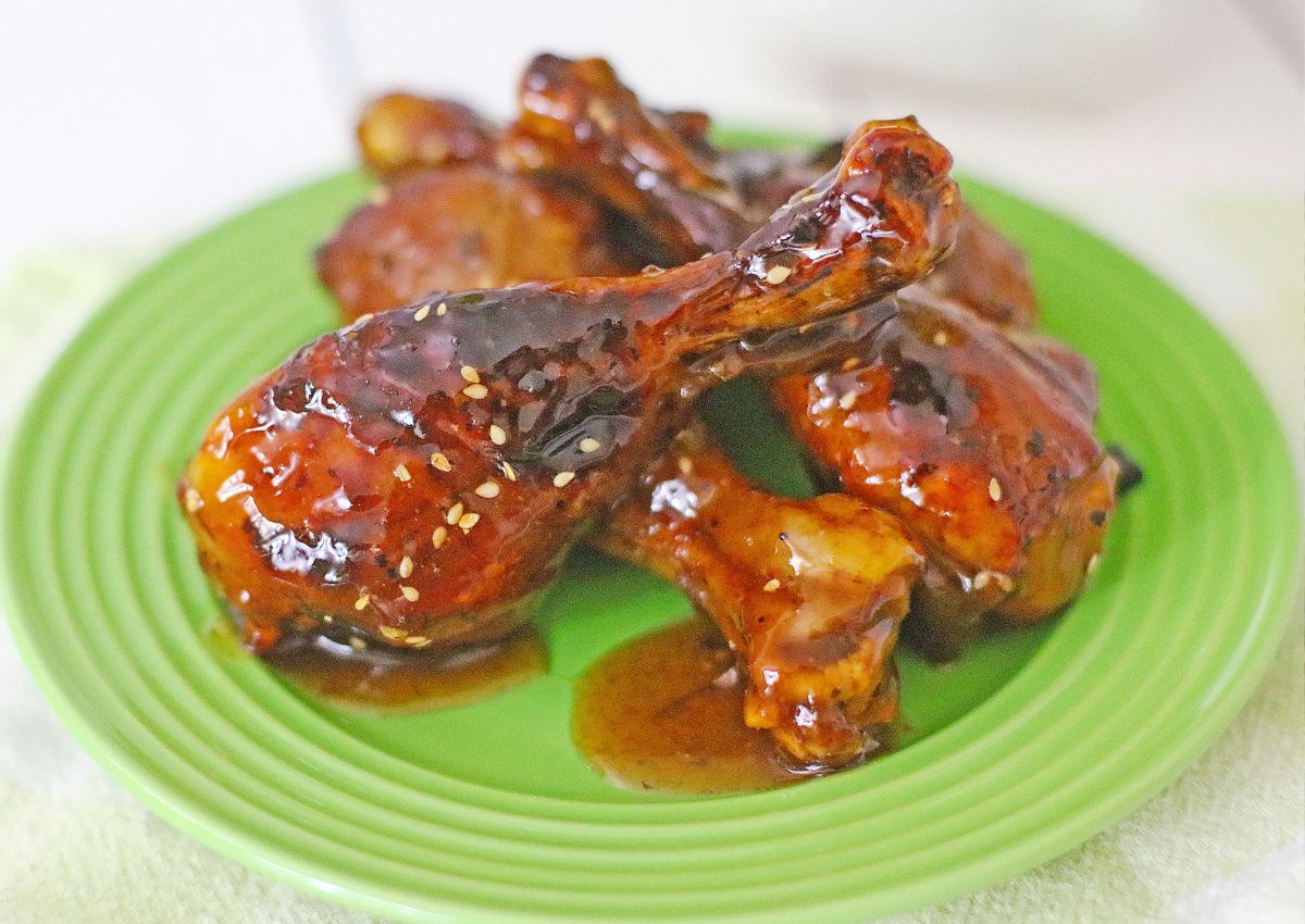Four glazed chicken drumsticks with a shiny, dark brown sauce and sesame seeds served on a green plate.