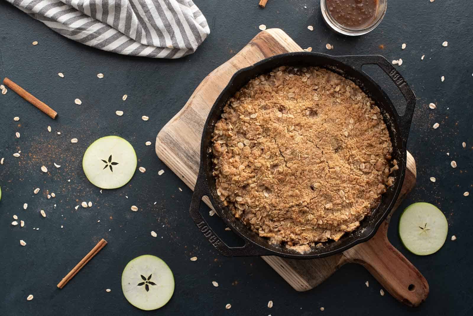 A cast iron skillet with apple crisp on a wooden board, surrounded by sliced green apples, oats, cinnamon sticks, and a striped cloth.