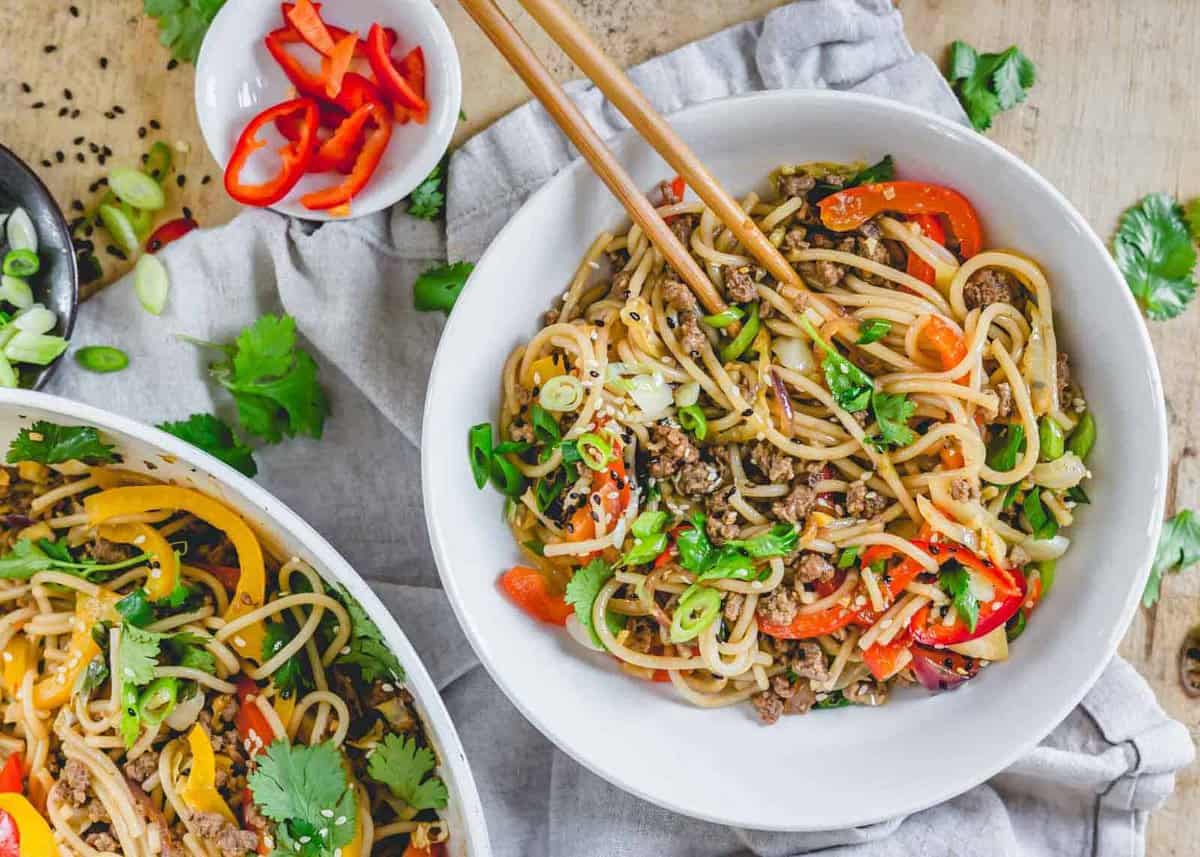 A bowl of stir-fried noodles with ground meat, sliced red and yellow bell peppers, scallions, and cilantro, with chopsticks resting on top. A side dish with sliced red peppers is nearby.