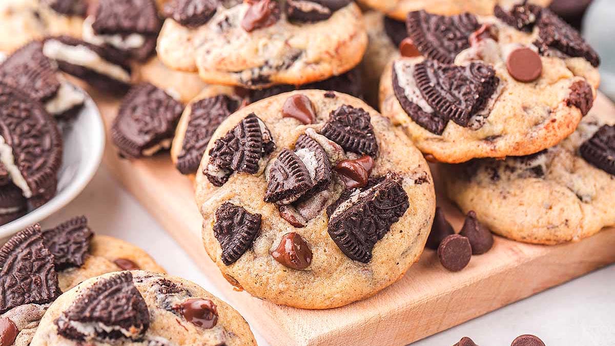 A close-up of chocolate chip cookies topped with large chunks of chocolate sandwich cookies on a wooden board, with extra cookie pieces on the side.
