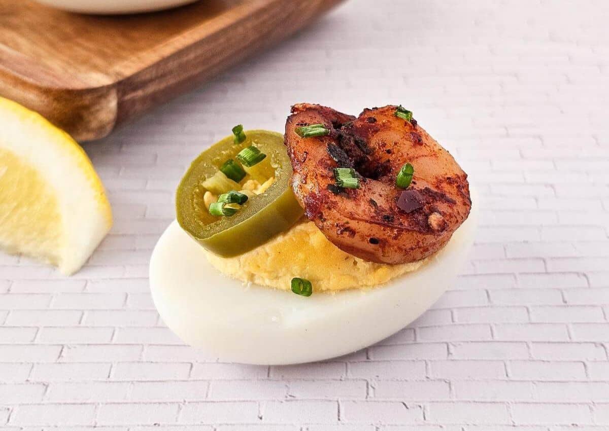 A deviled egg topped with a cooked shrimp, a jalapeño slice, and chopped chives sits on a white tiled surface with a lemon wedge nearby.