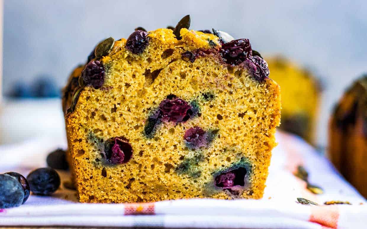 A close-up of a sliced blueberry loaf cake, showing its golden interior with visible berries and seeds on top, resting on a checkered cloth.