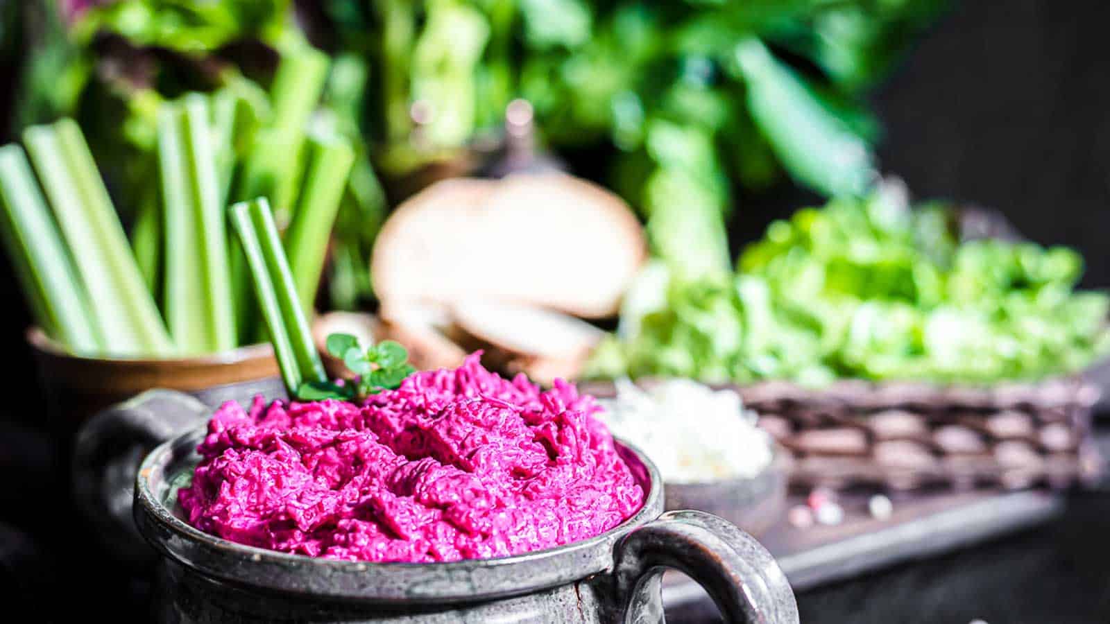 A bowl of vibrant purple beet dip garnished with herbs, surrounded by celery sticks and assorted fresh greens in the background.