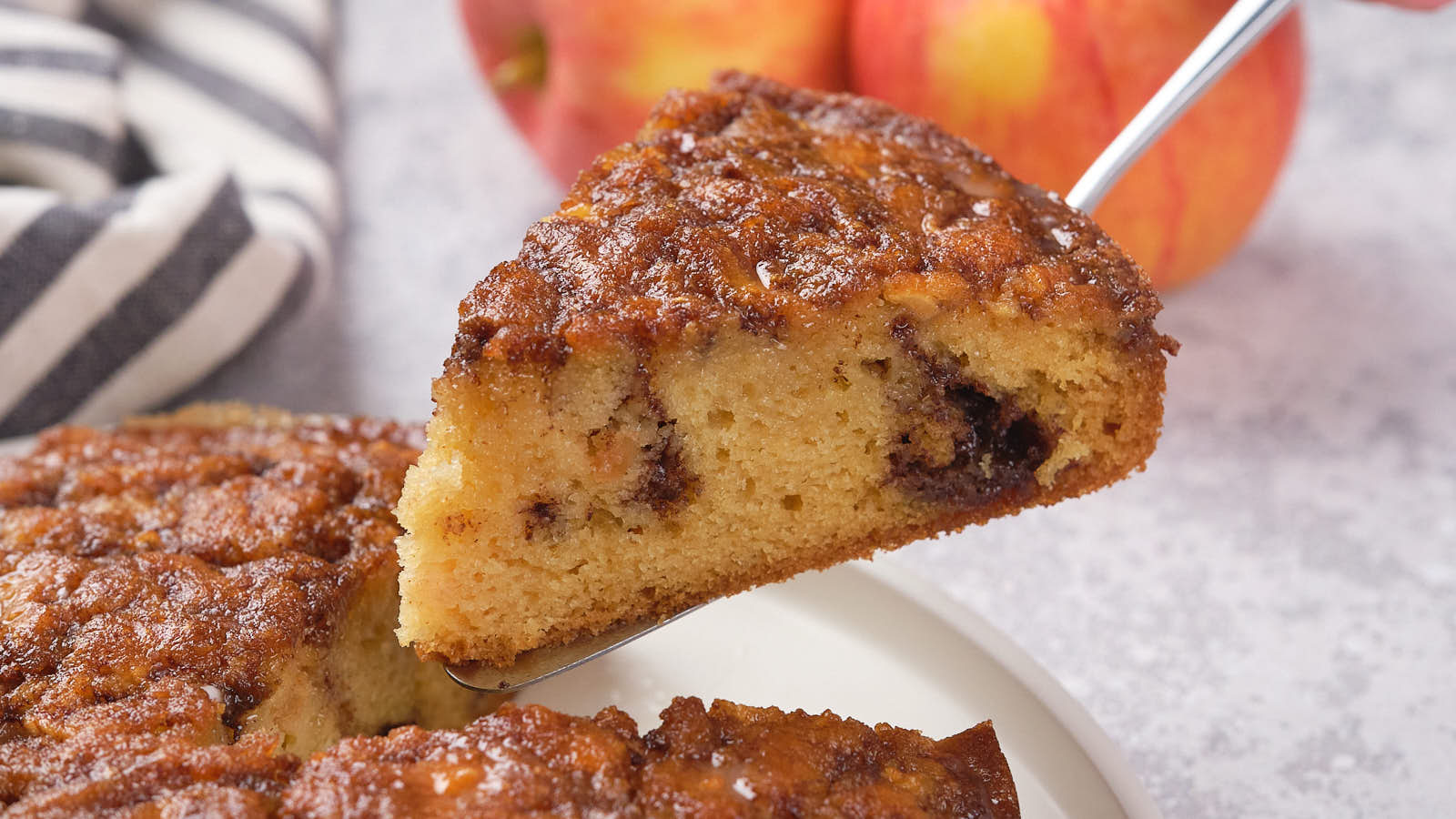 A slice of apple cake with visible cinnamon swirl being lifted from a whole cake, with apples and a striped cloth in the background.