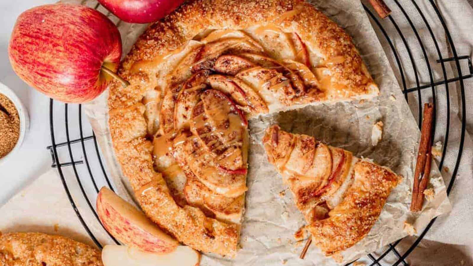 A rustic apple galette with one slice missing sits on parchment paper atop a wire rack, surrounded by fresh apples, cinnamon sticks, and apple slices.