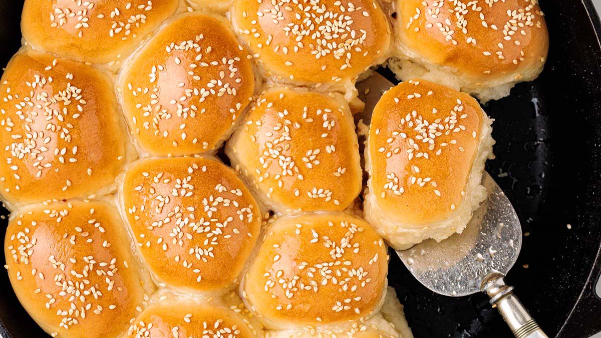 A tray of golden-brown dinner rolls topped with sesame seeds, with one roll being lifted out using a metal spatula.