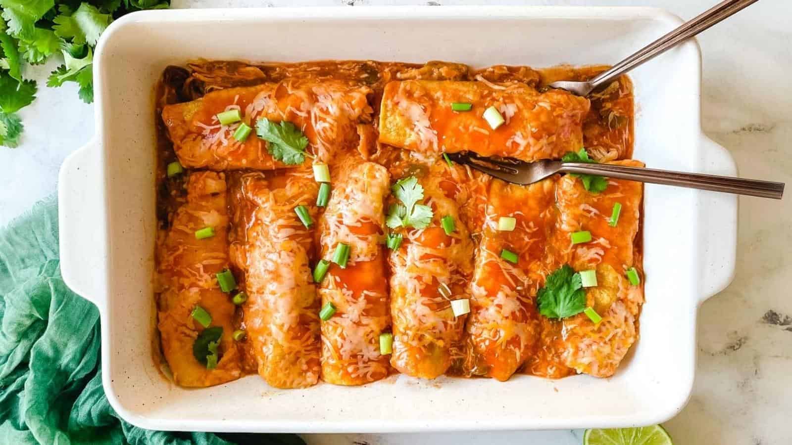 A white baking dish filled with six enchiladas topped with red sauce, melted cheese, chopped green onions, and cilantro, with two forks placed inside.