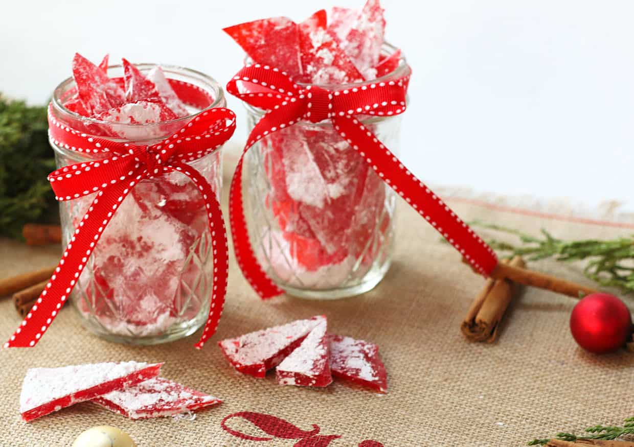 Two glass jars filled with red peppermint bark candy, tied with red ribbons, sit on a table with candy pieces, cinnamon sticks, and holiday decor.