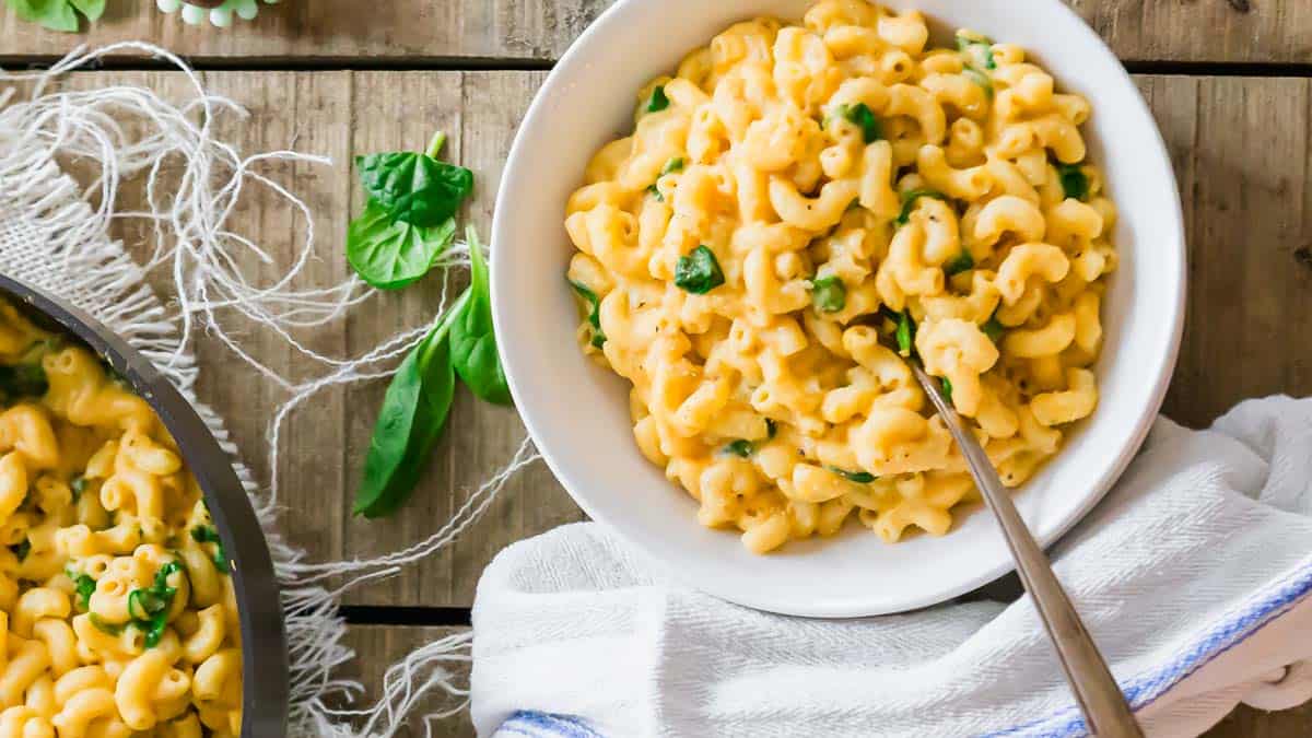 A bowl of macaroni and cheese with spinach, placed on a wooden table next to a pot and fresh spinach leaves.