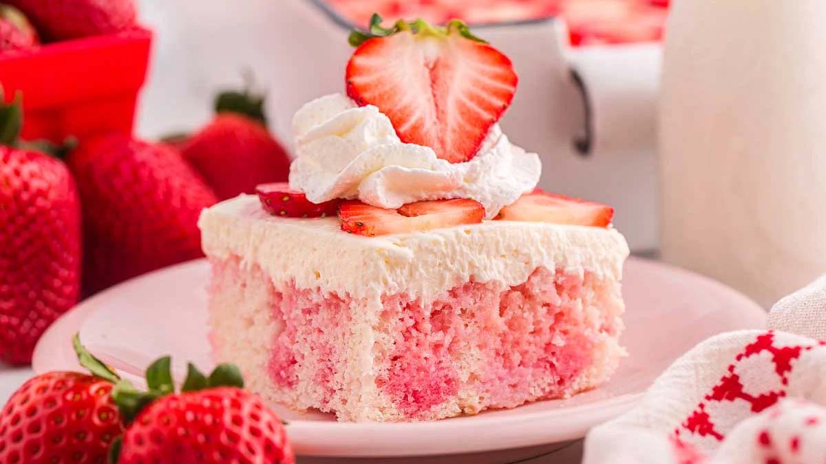 A slice of strawberry poke cake topped with whipped cream and sliced strawberries on a pink plate, surrounded by fresh strawberries.
