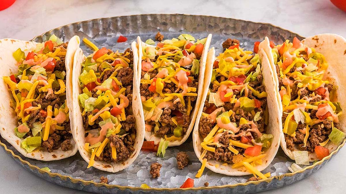 Four soft shell tacos filled with ground beef, shredded cheese, lettuce, diced tomatoes, and a drizzle of sauce, arranged on an oval serving tray.