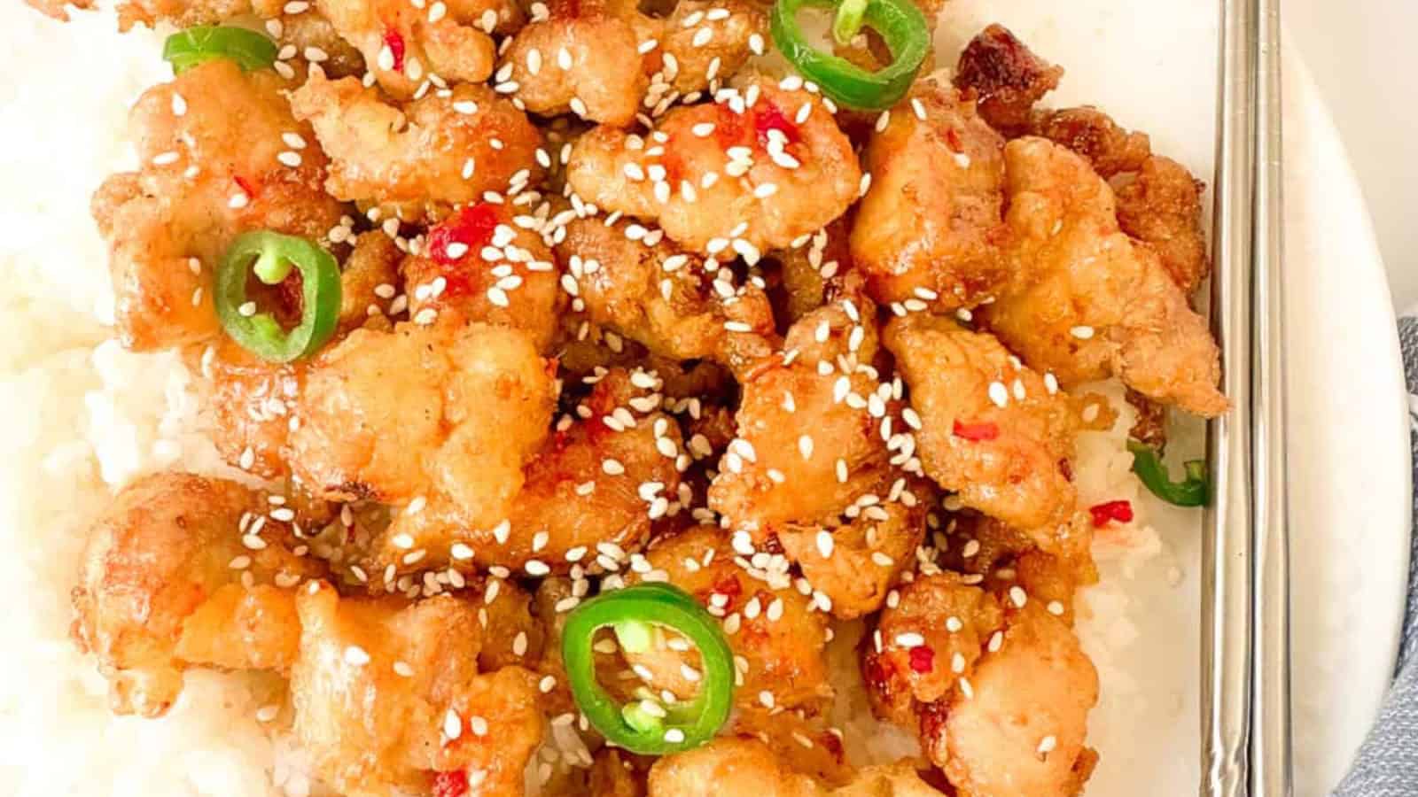 A plate of crispy fried chicken bites topped with sesame seeds and sliced green chili peppers, served over white rice.