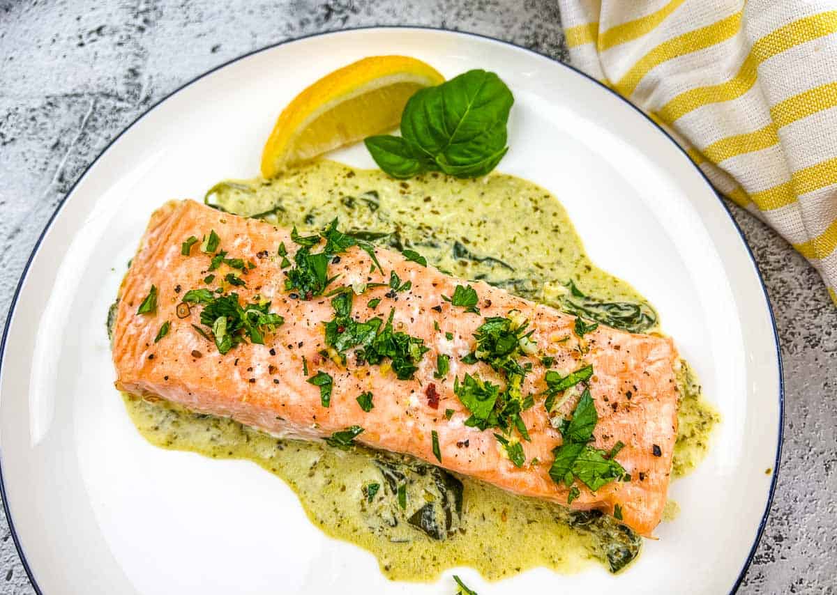 A cooked salmon fillet topped with chopped herbs sits on a creamy spinach sauce, garnished with a lemon wedge and basil leaf on a white plate.
