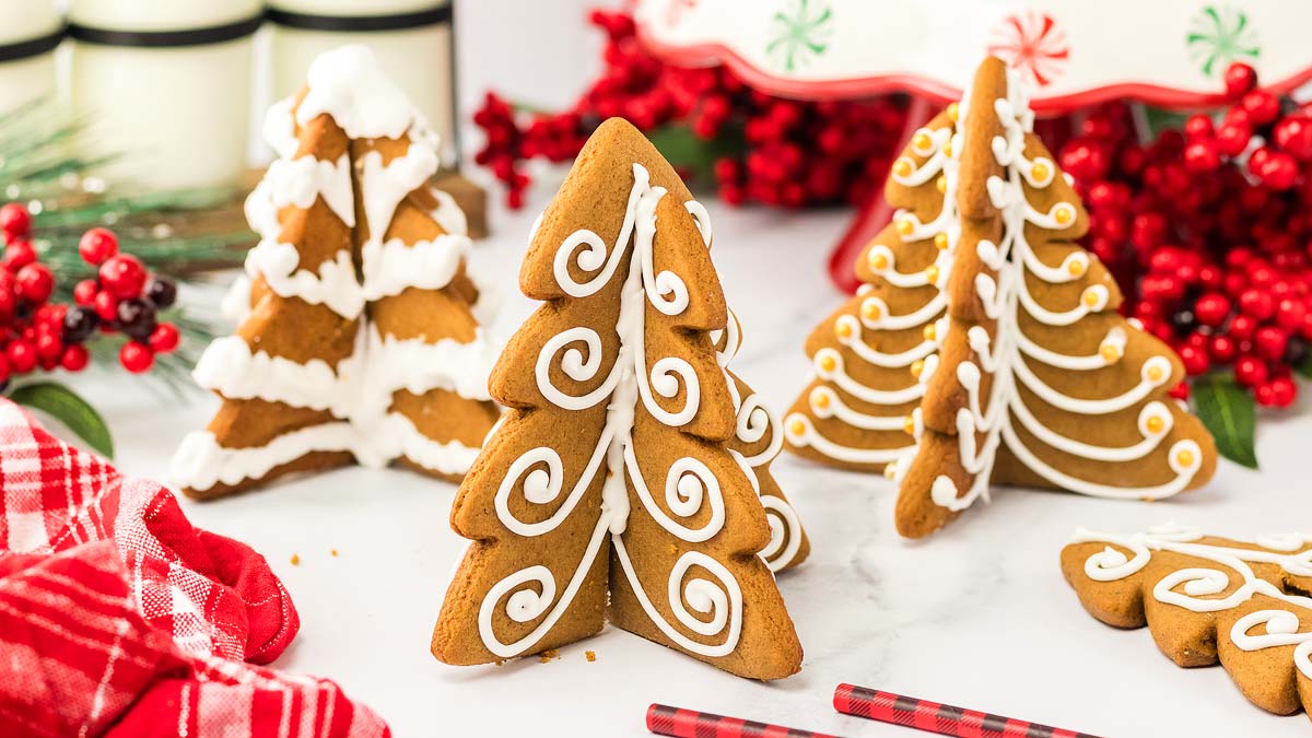 Three gingerbread cookies shaped like Christmas trees stand decorated with white icing. Red berries, a red plaid cloth, and tree-shaped cookies appear in the background.