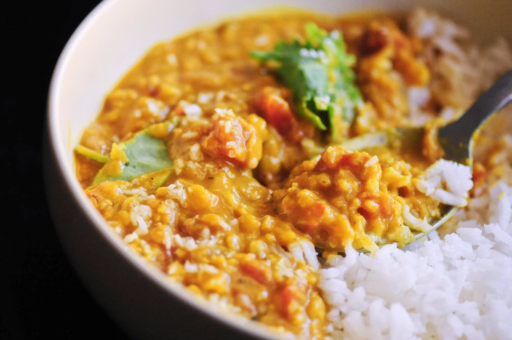 A close-up of a bowl containing creamy yellow lentil curry with cilantro, served alongside white rice, and a spoon scooping up the food.