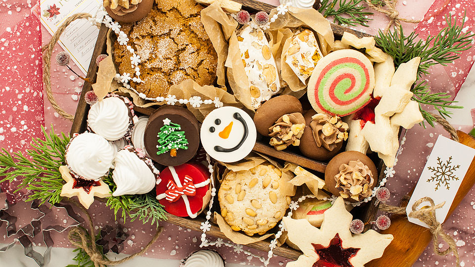A wooden tray filled with assorted decorated holiday cookies, meringues, and pastries, surrounded by festive greenery and tags.
