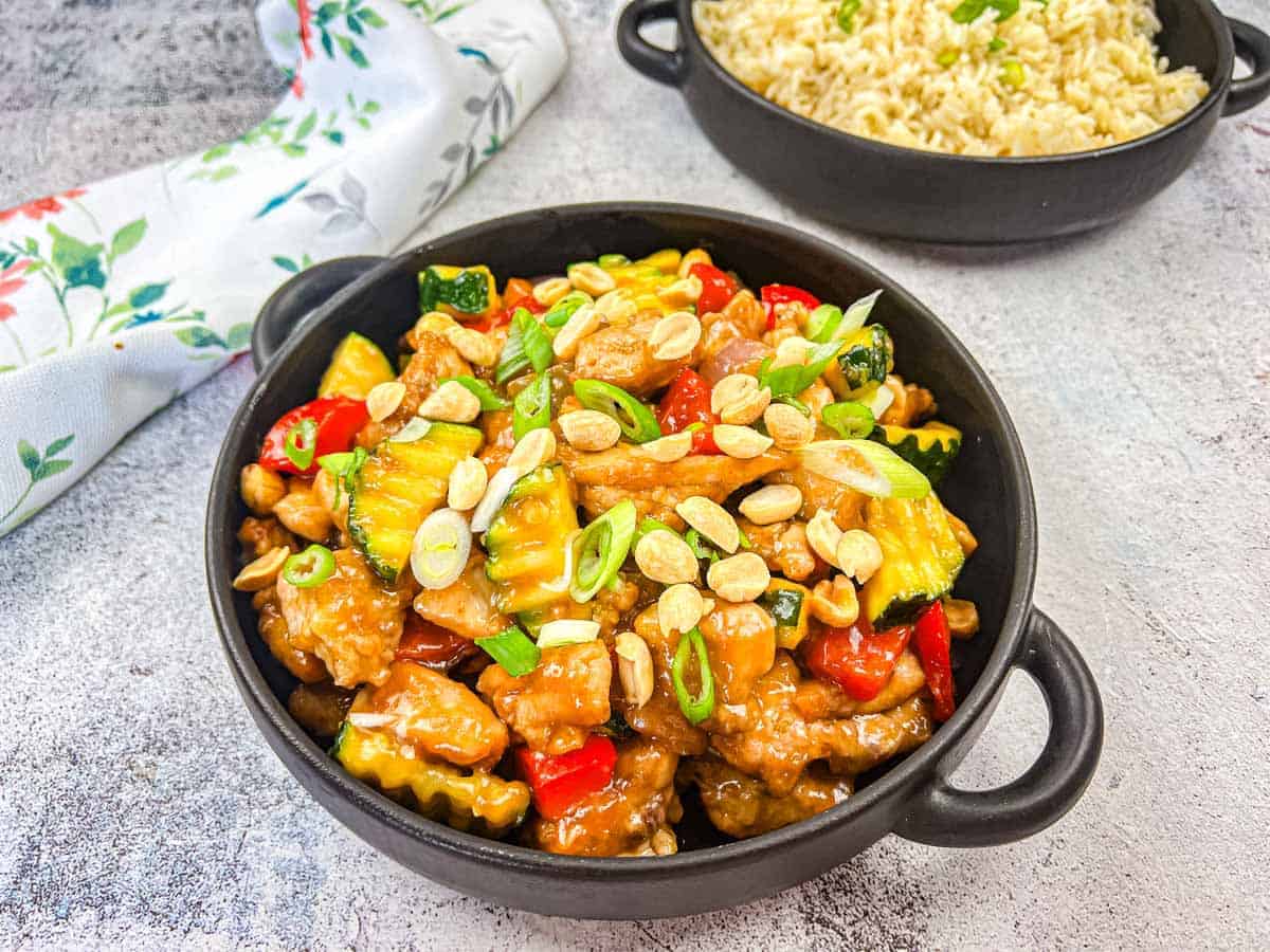 A black bowl filled with stir-fried chicken, vegetables, peanuts, and green onions sits next to a bowl of rice on a light surface.