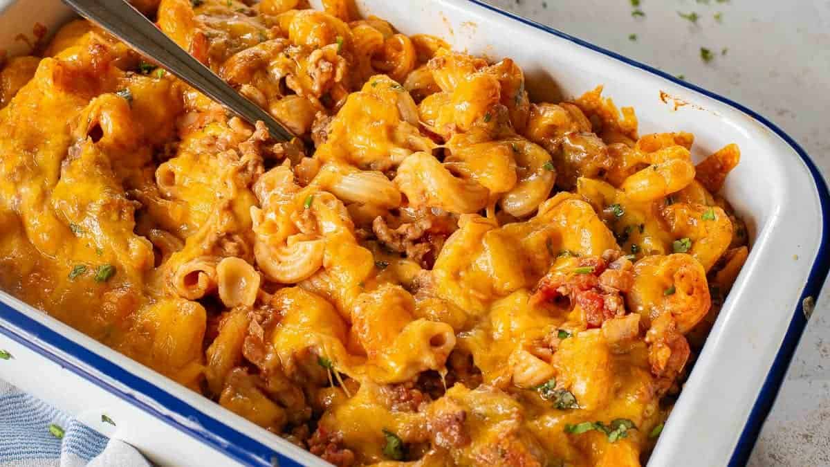 A baking dish filled with cheesy pasta casserole mixed with ground meat and topped with melted cheddar cheese. A serving spoon is partially inserted into the dish.