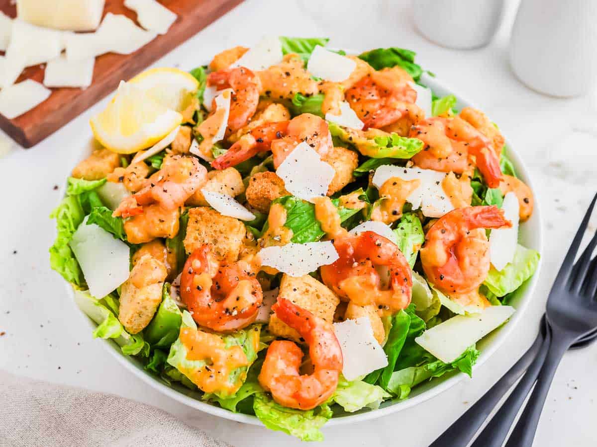 A bowl of Caesar salad topped with grilled shrimp, croutons, shaved Parmesan cheese, and Caesar dressing, with a lemon wedge on the side.