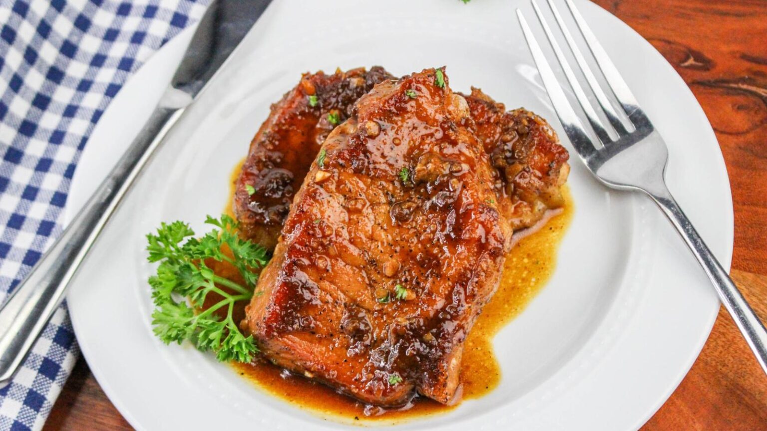 A white plate with glazed cooked pork chops garnished with parsley, next to a fork and knife on a blue checkered napkin.