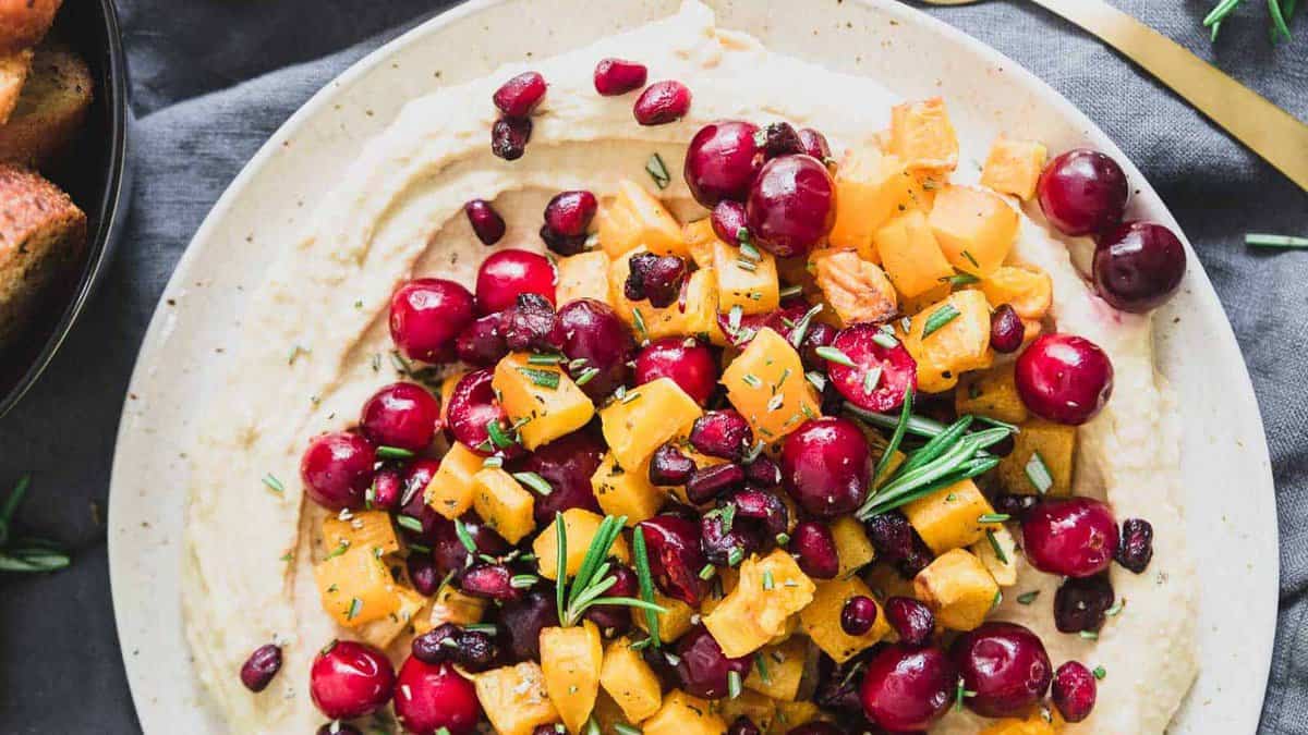 A plate of hummus topped with diced yellow squash, red grapes, pomegranate seeds, and rosemary sprigs.