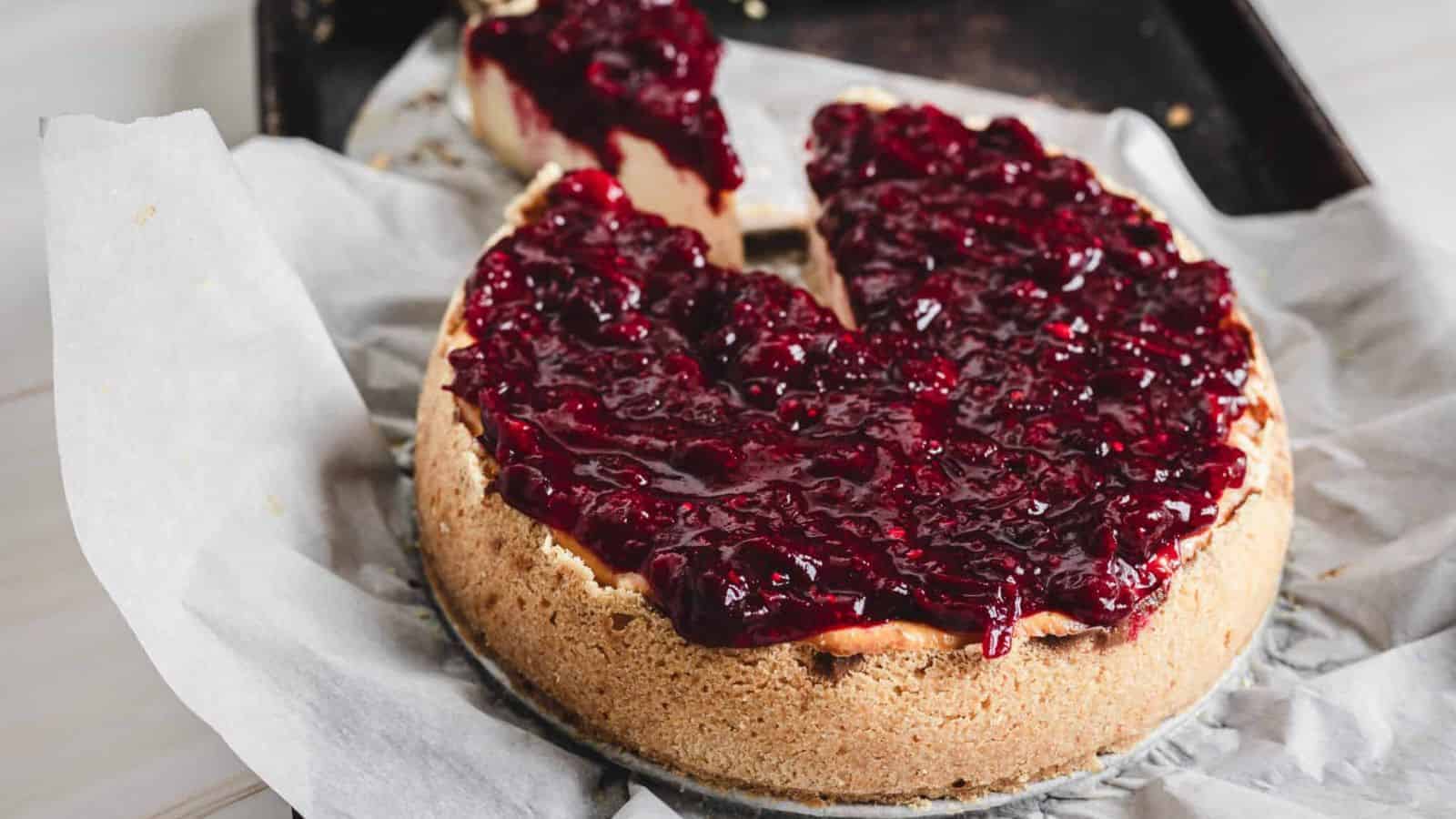 A cheesecake topped with a thick layer of berry compote sits on parchment paper, with one slice cut and slightly separated from the cake.