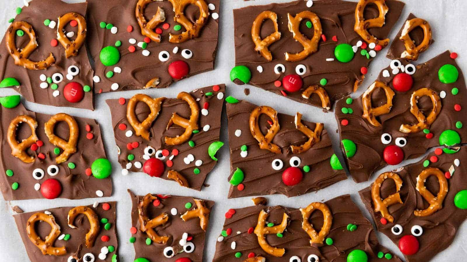 Chocolate bark pieces decorated with pretzels, red and green candies, and candy eyes to resemble reindeer faces, arranged on a white surface.