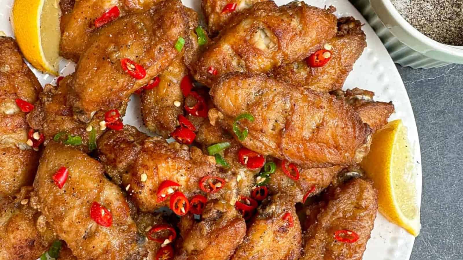 A plate of crispy fried chicken wings garnished with sliced red chilies, green onions, and lemon wedges, with a bowl of black pepper on the side.