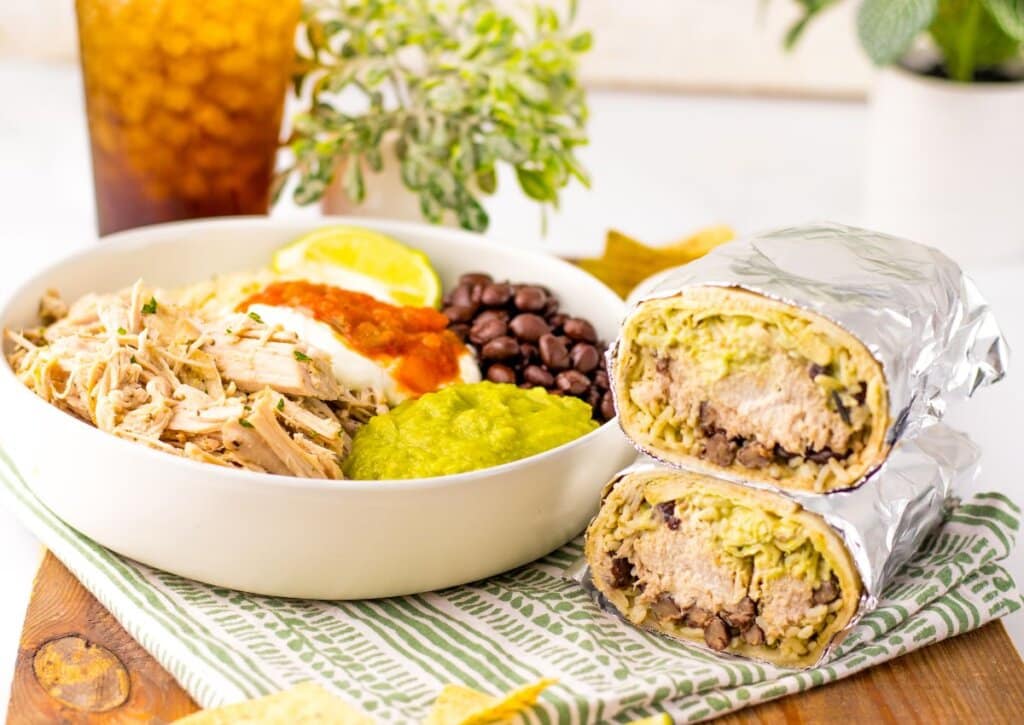 A bowl with shredded chicken, beans, salsa, and guacamole sits beside a halved burrito filled with rice, beans, and guacamole; iced drink in the background.
