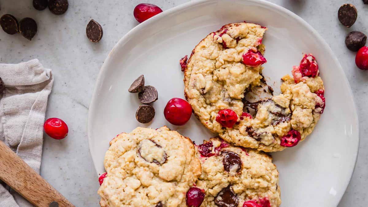 A white plate holds chunky cookies with dark chocolate pieces and red cranberries, surrounded by scattered chocolate chips and cranberries on a light surface.