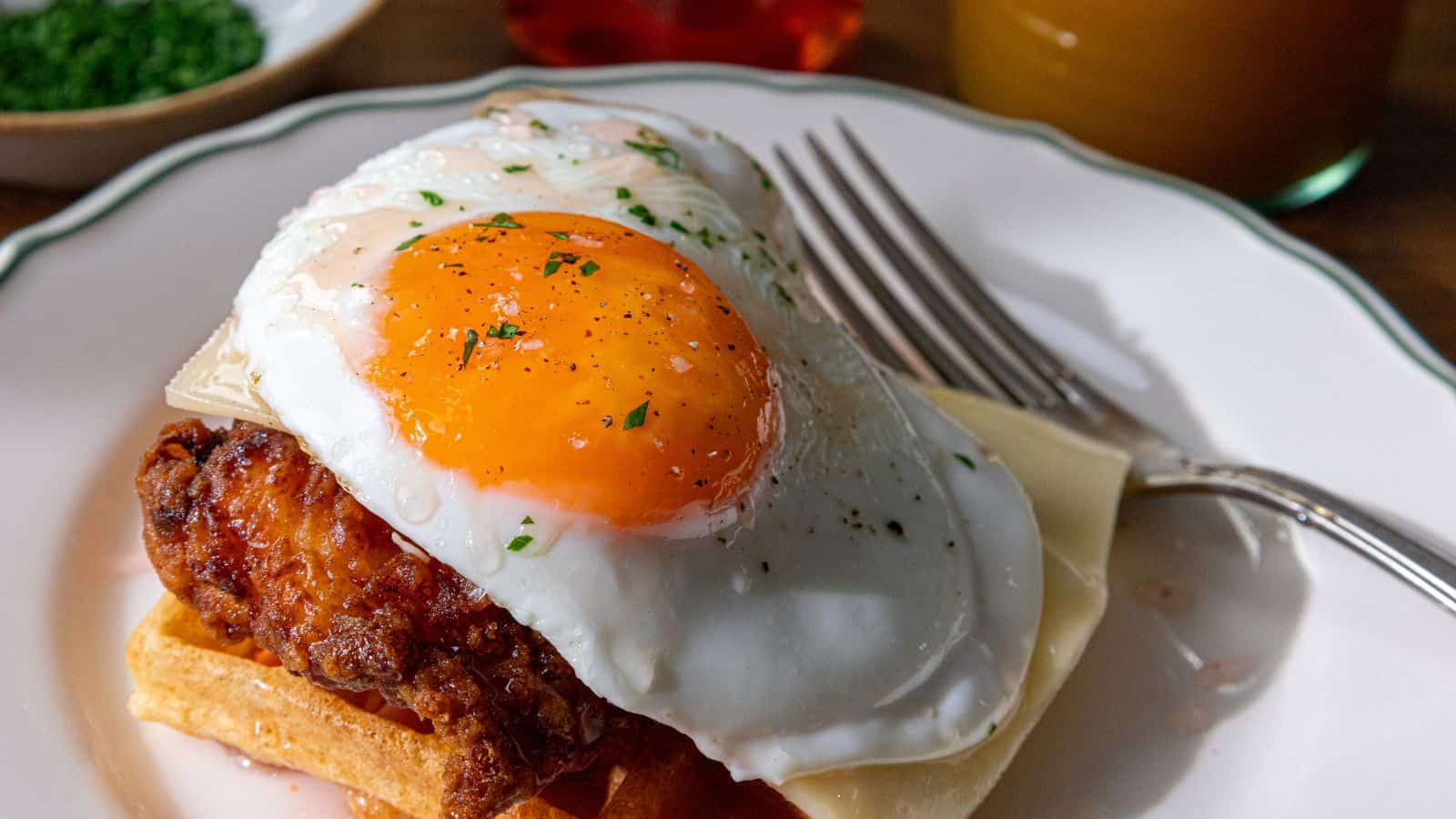A plate with a waffle topped with fried chicken, a slice of cheese, and a sunny-side-up egg, garnished with herbs, next to a fork.