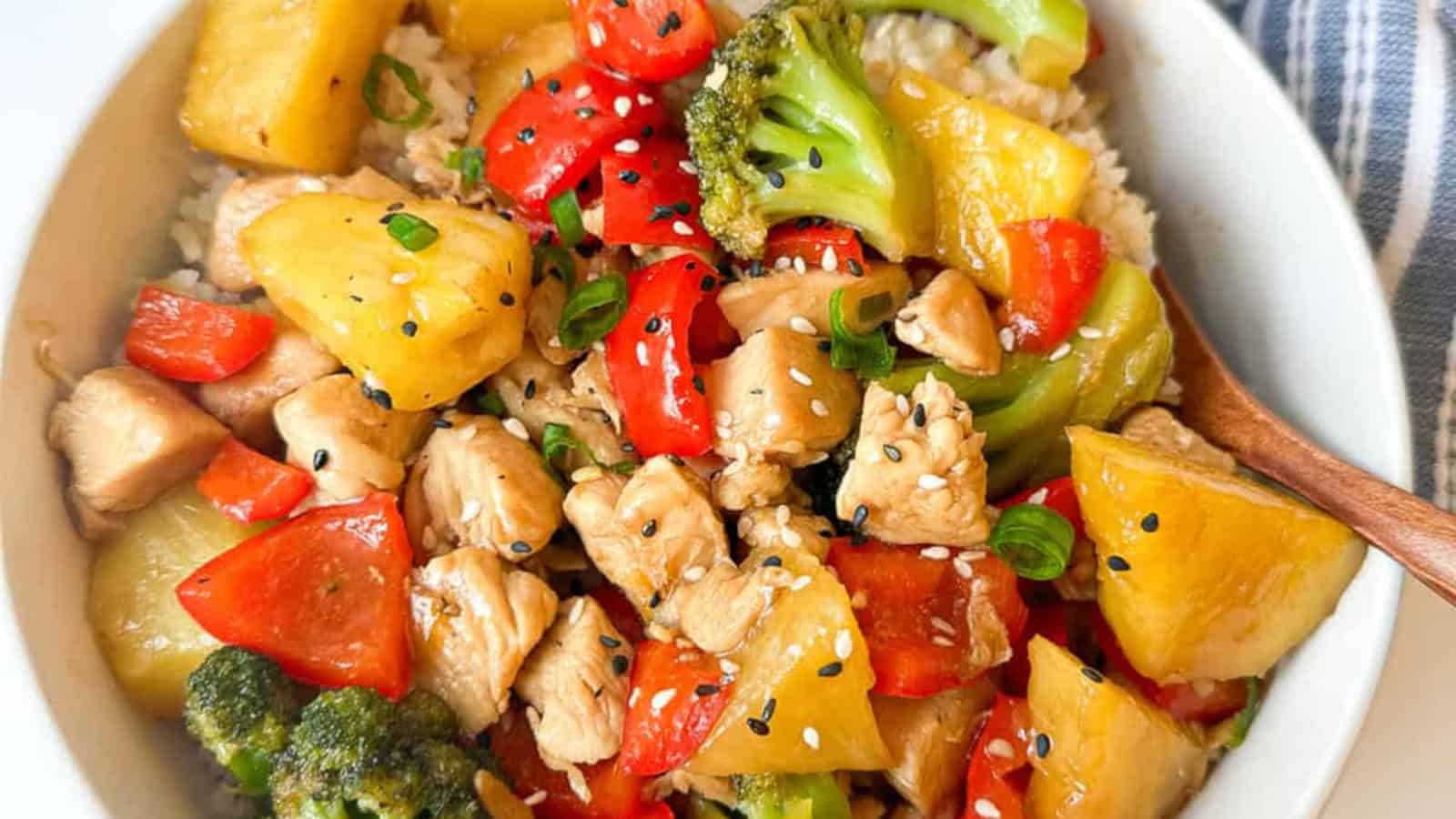 A bowl of stir-fried chicken with broccoli, red bell pepper, pineapple chunks, white rice, and sesame seeds, garnished with chopped green onions.