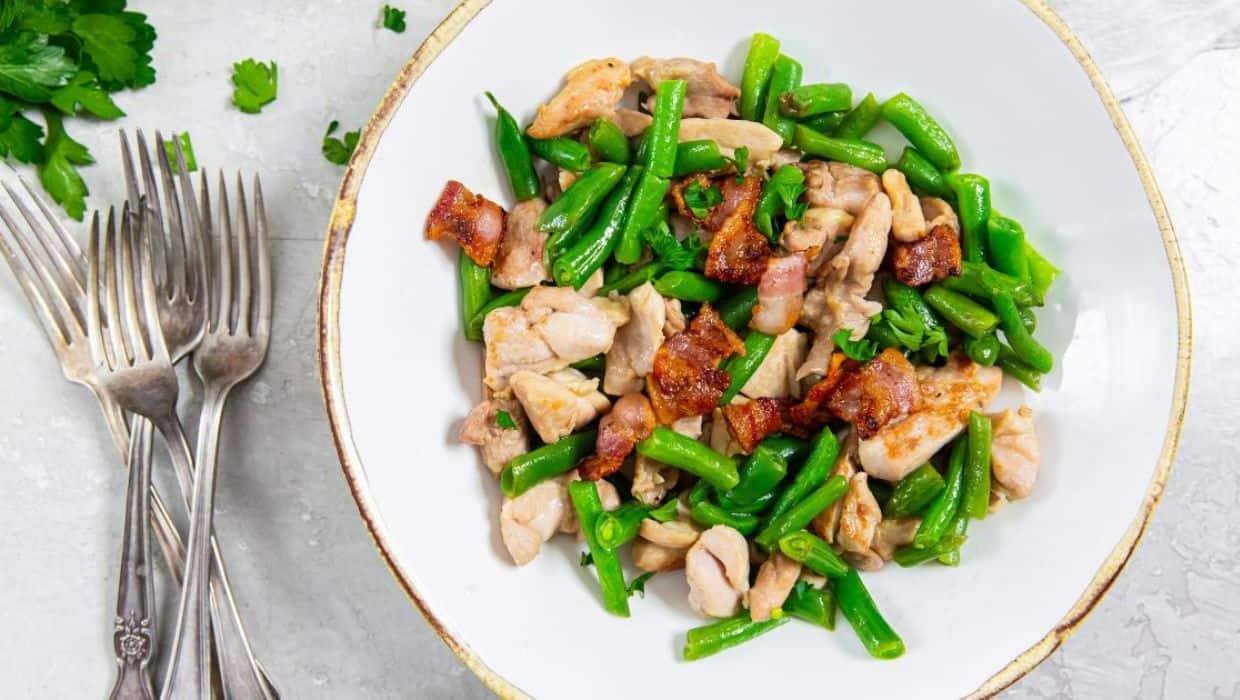 A white bowl filled with cooked green beans, pieces of chicken, and crispy bacon, with four forks beside the bowl on a light surface.
