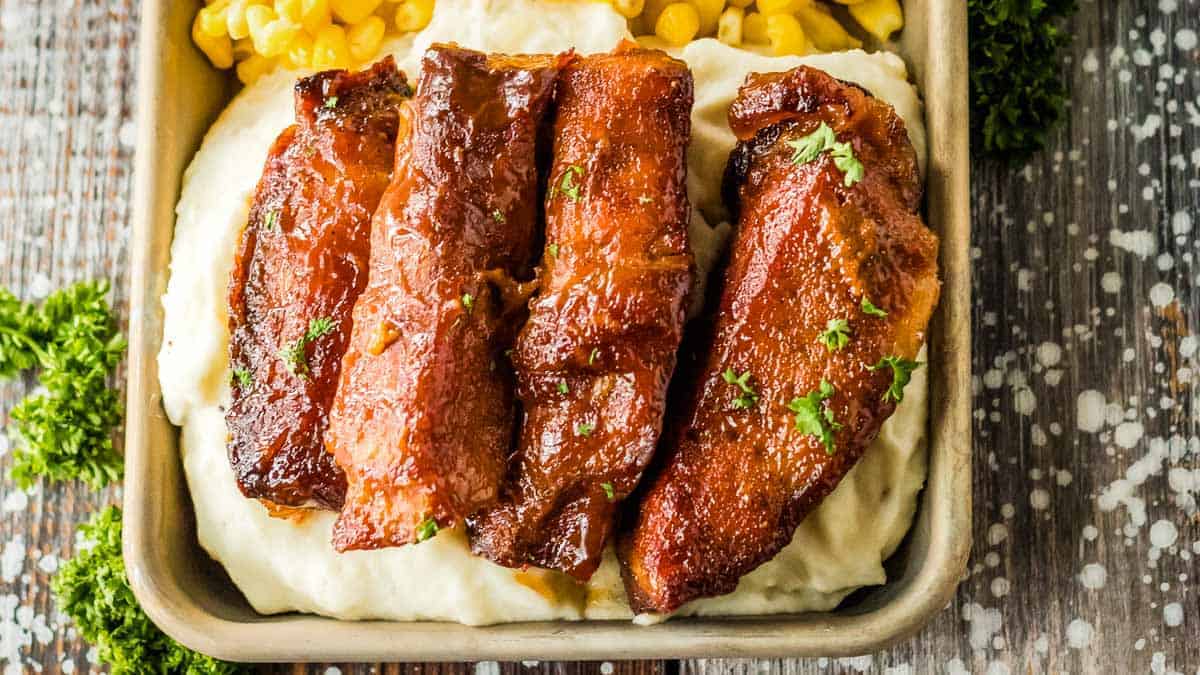 Four glazed pork ribs on a bed of mashed potatoes, served in a square dish with corn in the background.