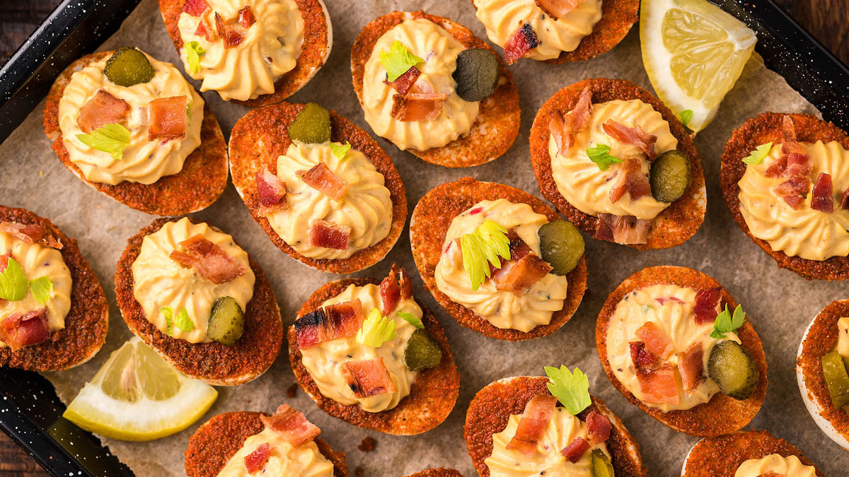 A tray of deviled eggs on crispy bases, topped with piped filling, bacon pieces, pickles, and herbs, with lemon wedges on the side.
